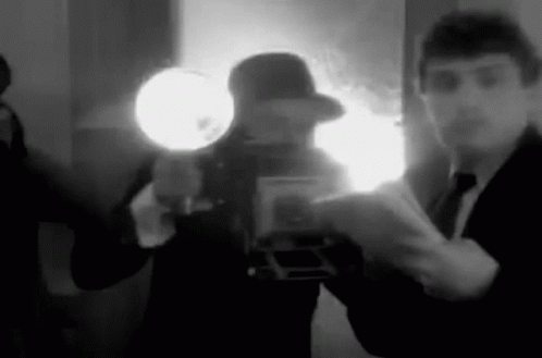 Black and white animated gif of a short video clip.  Reporter with pen and pad asking questions while several photographers with old school crown graphic or similar cameras with external big dish flash off the side are taking photos.