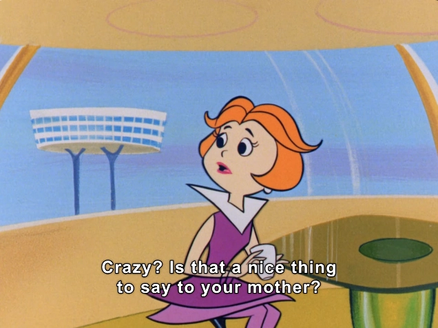 The image features a character with short, vibrant orange hair styled in a retro look, wearing a purple dress with a white collar. She has a surprised expression, with wide eyes and raised eyebrows, indicating a moment of disbelief or concern. The character appears to be situated in a futuristic interior, characterized by smooth lines and bright colors, including a rounded green table in front of her.

In the background, a stylized architectural structure can be seen through large, curved windows, enhancing the sci-fi ambiance. The scene includes dialogue text that reads, "Crazy? Is that a nice thing to say to your mother?", suggesting a comedic or dramatic context, typical of animated family interactions. The overall atmosphere is lively and playful, reminiscent of mid-20th century animated television shows.