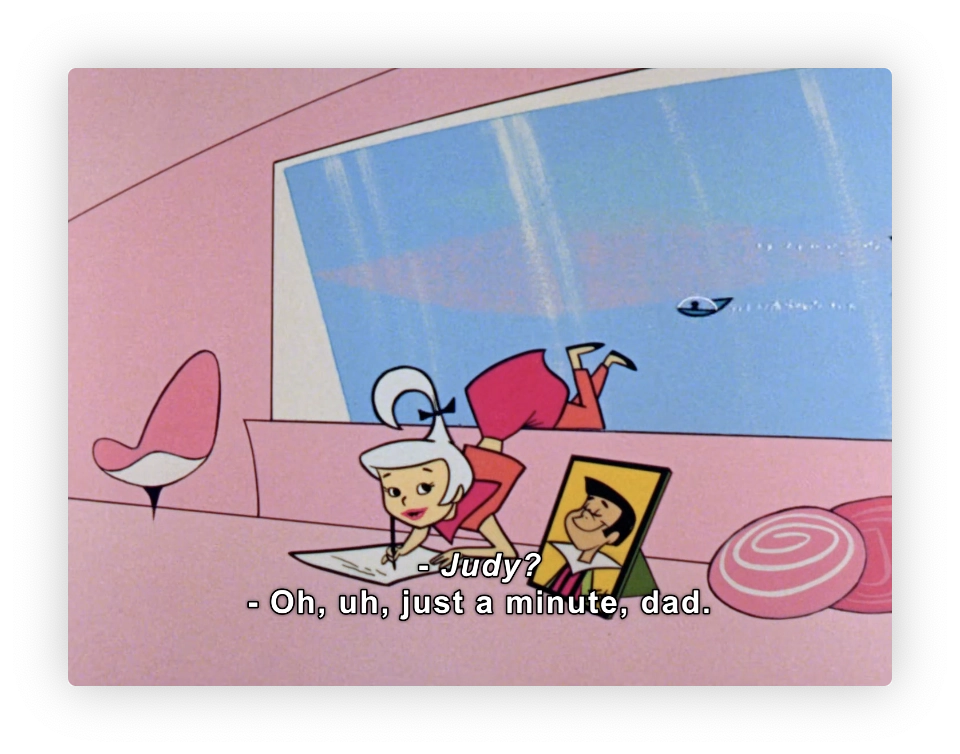 This image shows a scene from a cartoon. In it, there's a young female character sitting at a desk focused on her work. She appears to be distracted or calling out as she responds, "Oh, uh, just a minute, dad." The setting includes a large window that overlooks a futuristic landscape with a flying vehicle passing by. The interior has a retro-futuristic design with pastel colors and modernist furniture. This is indicative of mid-20th century animated representations of the future.
