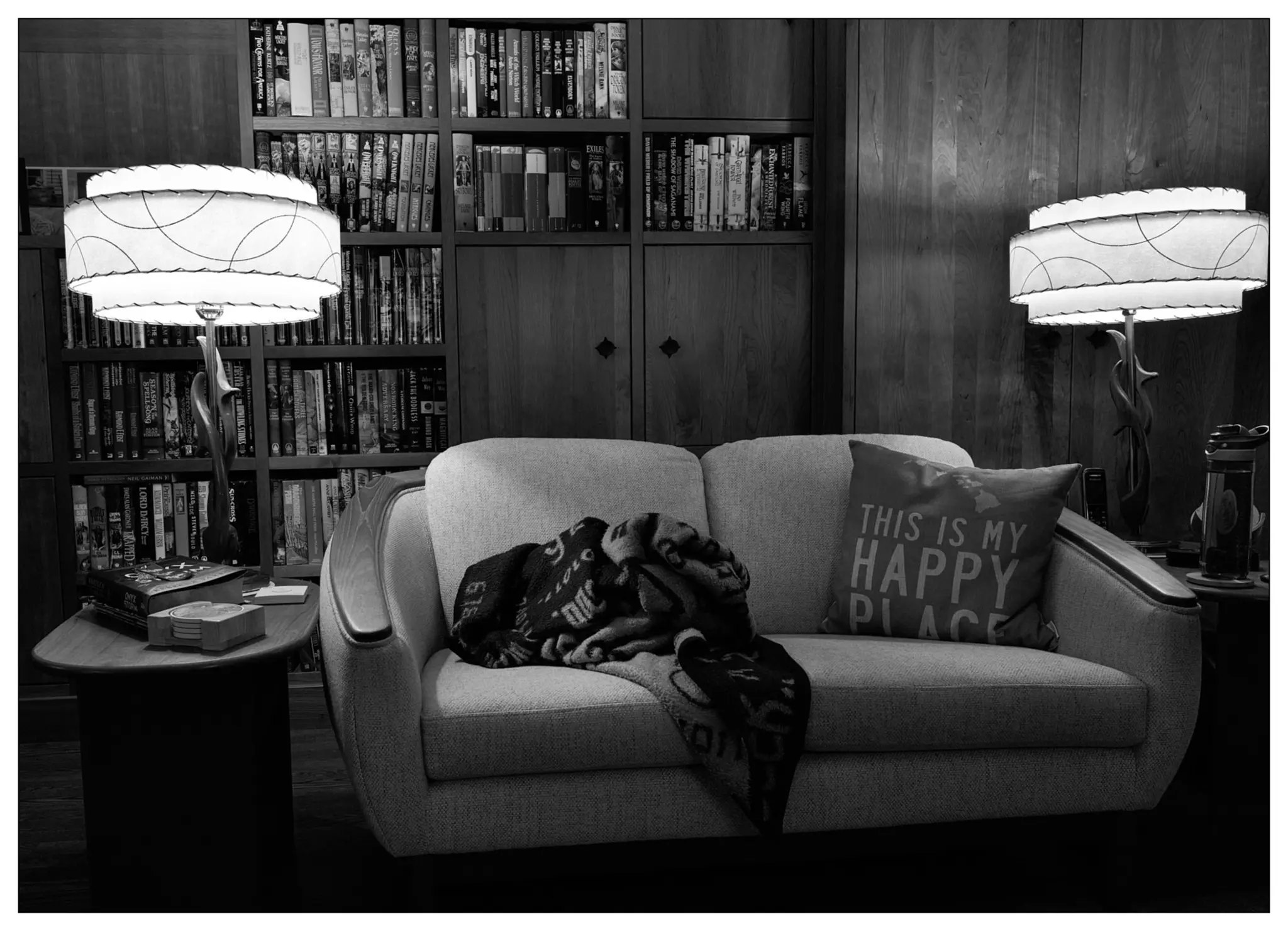 Black and white 5x7 image of a love seat (two seat couch) with end tables and lamps on either side.  There is a book case behind.  On the love seat is a rumpled blanket and a pillow that says "This is my happy place".