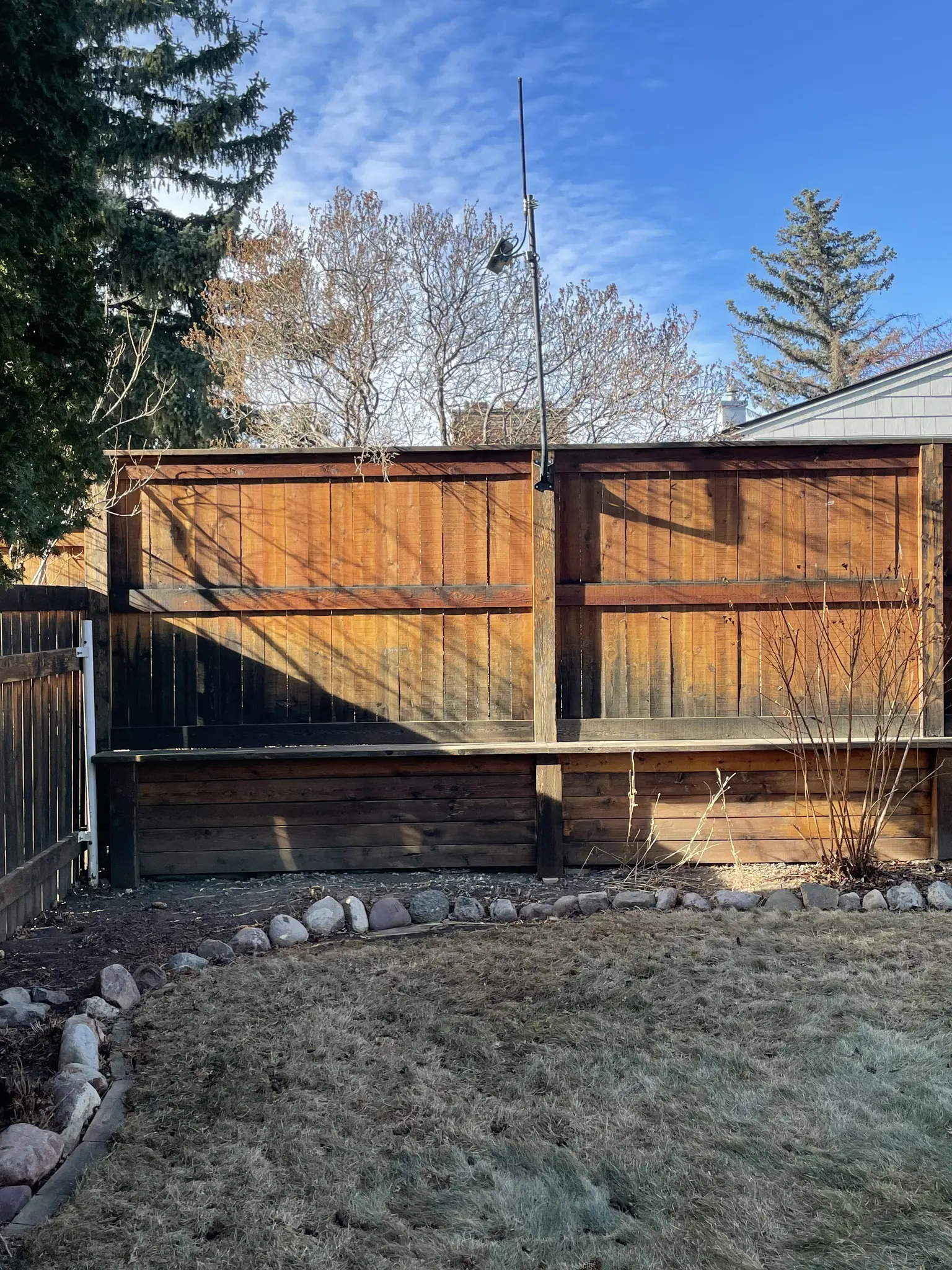 Portion of back yard with cedar fence, upon which is mounted a 5 foot pole with solar mesh node and meter or so tall antenna on top.   The sun is shining strong on the fence an hopefully the solar panel.