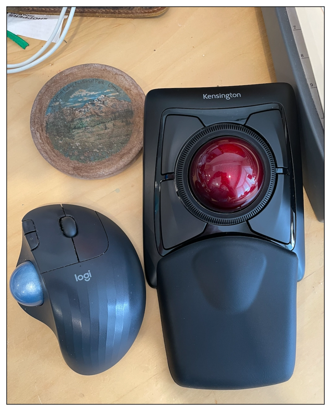 Overhead view of a desk, close up on a drink coaster and two 'mouse' devices.  One is a thumb track ball mouse, and another is a larger finger tip track ball.