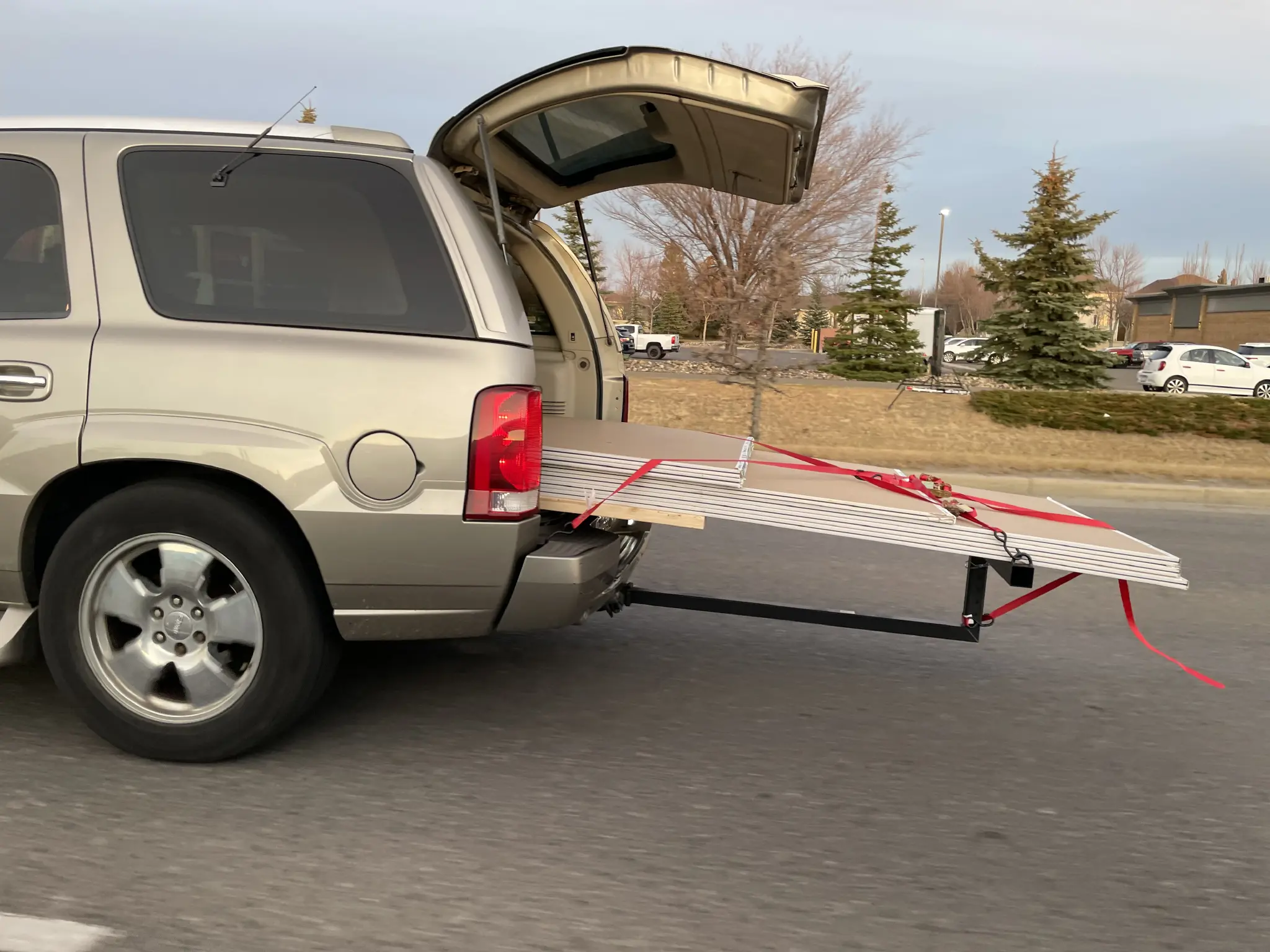 Side view.  SUV with rear hatch open and long poles of drywall hanging out.  A support is attached to the trailer hit hitch receiver that goes to a T holding end of drywall up.